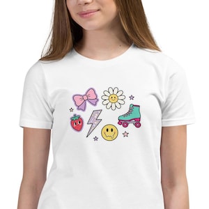 May include: White t-shirt featuring colorful cartoon graphics. The design includes a pink bow, daisy, strawberry, lightning bolt, roller skate, smiley face, and stars. The shirt is made of a soft material.