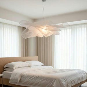Pode incluir: Uma luminária pendente grande e escultural com uma cúpula de tecido branco translúcido e em camadas que cria uma aparência suave semelhante a uma nuvem. A luz é suspensa no teto por um fio fino, emitindo um brilho quente.