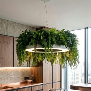 May include: A circular white pendant light fitting suspended from the ceiling, featuring a thick ring design with an integrated LED light source and a dense arrangement of lush green artificial foliage cascading from the top and sides.