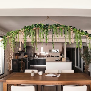 Puede incluir: Una mesa de comedor de madera con cuatro sillas blancas. Encima de la mesa hay un arco decorativo de madera y cuerda, adornado con plantas verdes colgantes. La cocina es visible en el fondo.