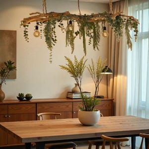 Op de afbeelding: Een rustieke houten kroonluchter met hangend groen en drie Edison-stijl gloeilampen verlicht een eetkamer. Een houten eettafel en stoelen, een plant in een pot, een houten kast en een raam met transparante gordijnen zijn te zien. De tafel is ongeveer 180 cm lang.