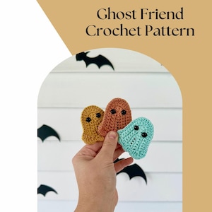 Könnte beinhalten: Drei gehäkelte Geister-Amigurumi-Spielzeuge in den Farben Orange, Gelb und Türkis. Jeder Geist hat zwei schwarze Knopfaugen. Die Geister werden in einer Hand vor einem weißen Holzhintergrund mit schwarzen Fledermäusen gehalten.