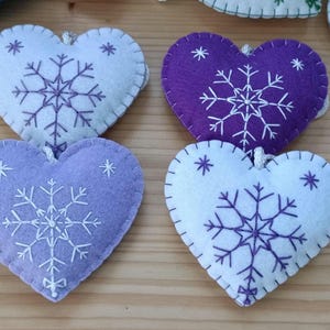 Close-up of four heart-shaped felt ornaments in shades of purple and white. Each ornament features a white embroidered snowflake design and a stitched border. A small loop is attached for hanging.