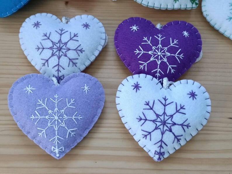 Close-up of four heart-shaped felt ornaments in shades of purple and white. Each ornament features a white embroidered snowflake design and a stitched border. A small loop is attached for hanging.