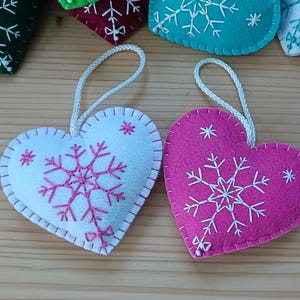Heart-shaped felt ornaments with snowflake embroidery. One is white with pink stitching, the other is pink with white stitching. Each has a white cord for hanging. Other ornaments are visible in the background.