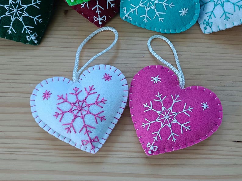Heart-shaped felt ornaments with snowflake embroidery. One is white with pink stitching, the other is pink with white stitching. Each has a white cord for hanging. Other ornaments are visible in the background.