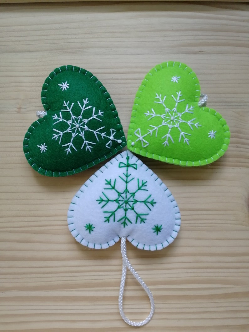 3 green and white christmas tree ornaments. all are felt hearts with hand embroidered snowflake. snowflake is in the center of the heart, and asides are two sparkles. snowflake is white on green hearts, and green on white heart, ends with a ribbon.
