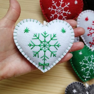 felt christmas tree ornament or winter festive decoration, felt heart held on a palm of a hand for size perception. heart is plush and with string. snowflake in its center is hand embroidered, and has embroidered ribbon at bottom. white and green.