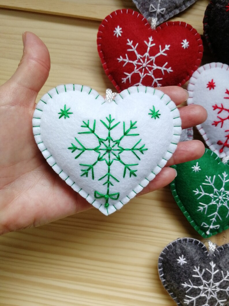 felt christmas tree ornament or winter festive decoration, felt heart held on a palm of a hand for size perception. heart is plush and with string. snowflake in its center is hand embroidered, and has embroidered ribbon at bottom. white and green.
