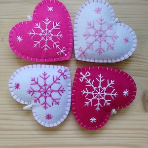 4 pink and white christmas tree ornaments. all are felt hearts with hand embroidered snowflake. snowflake is in the center of the heart, and asides are two sparkles. snowflake is white on pink hearts, and pink on white hearts, ends with a ribbon.