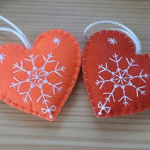 Two heart-shaped ornaments. Each ornament is made of orange felt and features a white embroidered snowflake design. A white cord is attached for hanging. The ornaments are suitable for holiday or seasonal decor.