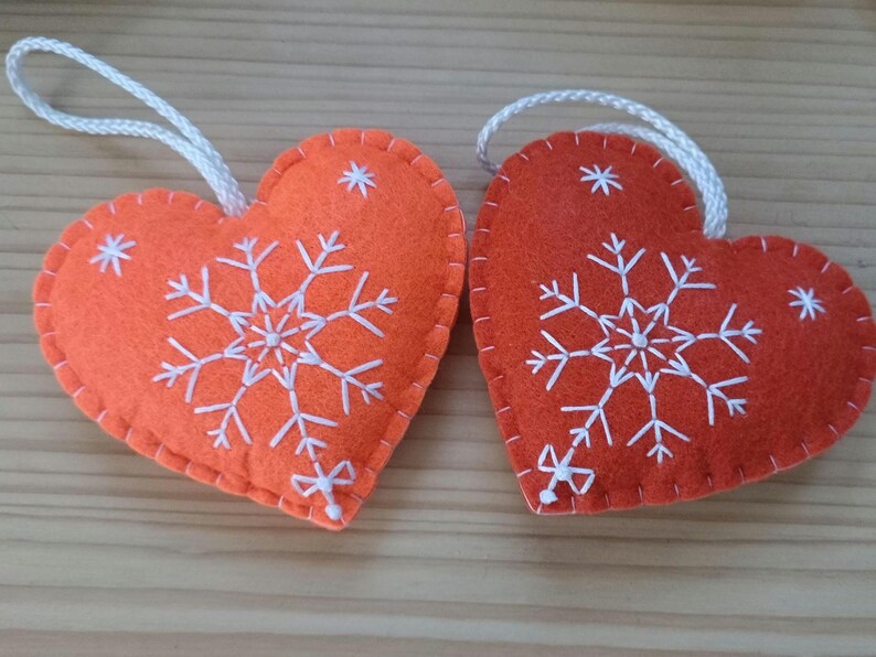 Two heart-shaped ornaments. Each ornament is made of orange felt and features a white embroidered snowflake design. A white cord is attached for hanging. The ornaments are suitable for holiday or seasonal decor.