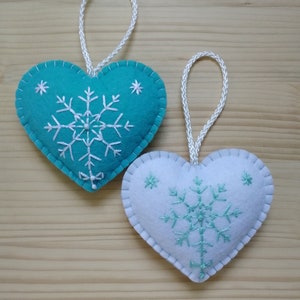 mint blue and white christmas tree ornaments. both are felt hearts with hand embroidered snowflake. snowflake is in the center of the heart, and asides are two sparkles. snowflake is white on blue heart, and blue on white heart, ends with a ribbon.