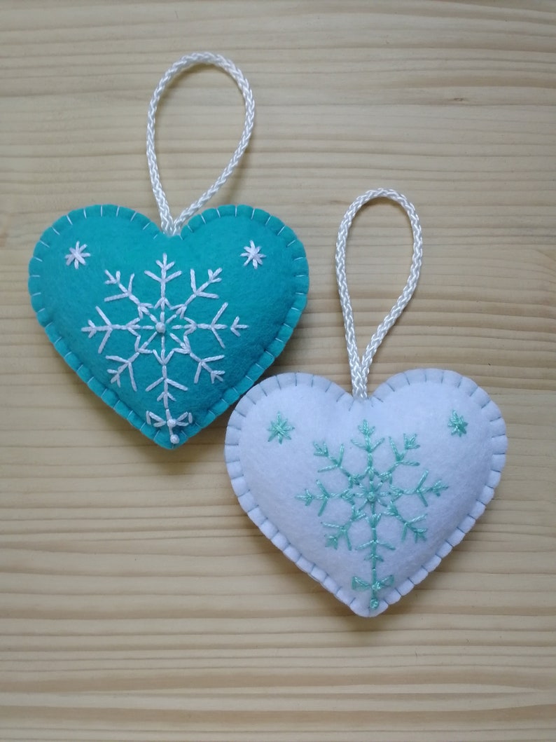 mint blue and white christmas tree ornaments. both are felt hearts with hand embroidered snowflake. snowflake is in the center of the heart, and asides are two sparkles. snowflake is white on blue heart, and blue on white heart, ends with a ribbon.