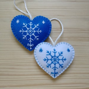 royal blue and white christmas tree ornaments. both are felt hearts with hand embroidered snowflake. snowflake is in the center of the heart, and asides are two sparkles. snowflake is white on blue heart, and blue on white heart. Ends with a ribbon.