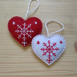 red and white christmas tree ornaments. both are felt hearts with hand embroidered snowflake. snowflake is in the center of the heart, and asides are two sparkles. snowflake is white on red heart, and red on white heart, and it ends with a ribbon.