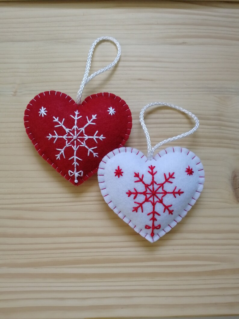 red and white christmas tree ornaments. both are felt hearts with hand embroidered snowflake. snowflake is in the center of the heart, and asides are two sparkles. snowflake is white on red heart, and red on white heart, and it ends with a ribbon.