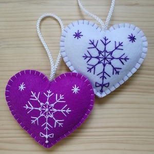 purple and white christmas tree ornaments. both are felt hearts with hand embroidered snowflake. snowflake is in the center of the heart, and asides are two sparkles. snowflake is white on purple heart, and purple on white heart, ends with a ribbon.