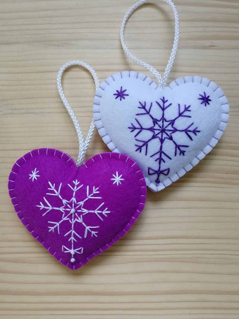 purple and white christmas tree ornaments. both are felt hearts with hand embroidered snowflake. snowflake is in the center of the heart, and asides are two sparkles. snowflake is white on purple heart, and purple on white heart, ends with a ribbon.