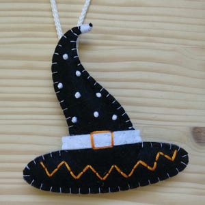 May include: A black felt witch hat ornament featuring white polka dots, a white band with an orange square buckle, and an orange zigzag stitch pattern along the brim. The hat is attached to a white string for hanging, with visible white stitching.