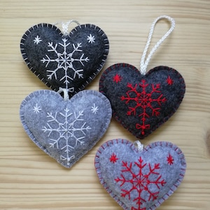 4 gray christmas tree ornaments in two shades. all are felt hearts with hand embroidered snowflake. snowflake is in the center of the heart, and asides are two sparkles. snowflake is either white or red on both gray shades, ends with a ribbon.