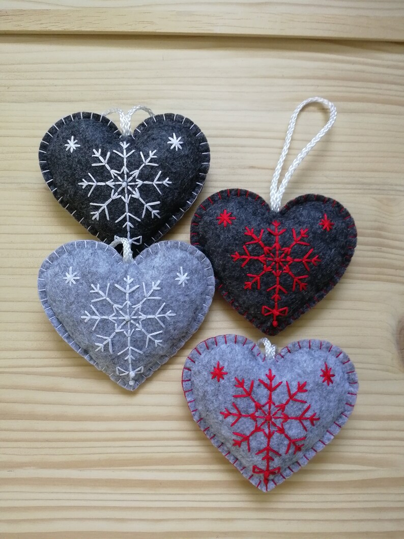 4 gray christmas tree ornaments in two shades. all are felt hearts with hand embroidered snowflake. snowflake is in the center of the heart, and asides are two sparkles. snowflake is either white or red on both gray shades, ends with a ribbon.