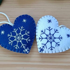 Two heart-shaped Christmas ornaments. One is navy blue, the other is white. Both have a snowflake design in the center and smaller star accents. Each ornament has a white string for hanging.