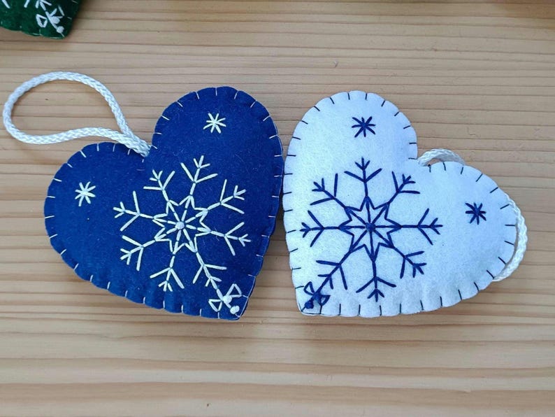 Two heart-shaped Christmas ornaments. One is navy blue, the other is white. Both have a snowflake design in the center and smaller star accents. Each ornament has a white string for hanging.