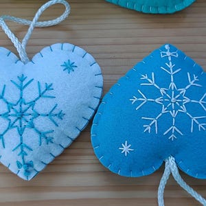 Two heart-shaped felt ornaments. One is white with a turquoise snowflake design and the other is blue with a white snowflake design. Both have white string for hanging. These ornaments are perfect for winter holiday decor.