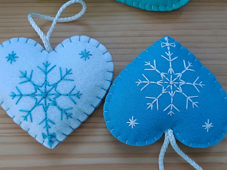 Two heart-shaped felt ornaments. One is white with a turquoise snowflake design and the other is blue with a white snowflake design. Both have white string for hanging. These ornaments are perfect for winter holiday decor.