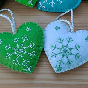 Close-up of two heart-shaped Christmas ornaments. One is green, the other white, both with white and green embroidered snowflake designs. Each ornament has a white string for hanging. The ornaments are made of felt.