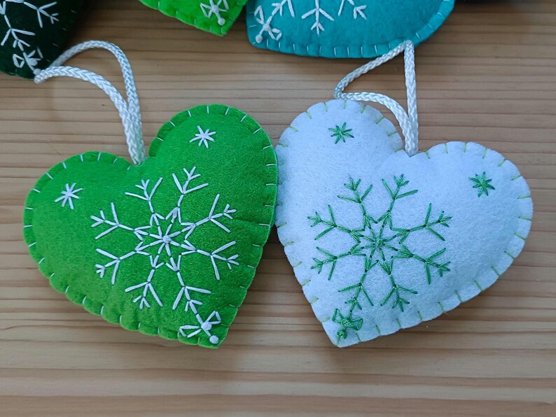 Close-up of two heart-shaped Christmas ornaments. One is green, the other white, both with white and green embroidered snowflake designs. Each ornament has a white string for hanging. The ornaments are made of felt.
