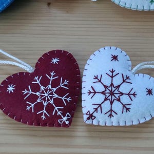 Two heart-shaped Christmas ornaments. One is burgundy with white snowflake embroidery, the other is white with burgundy snowflake embroidery. Both have white string loops for hanging. The ornaments are made of felt.