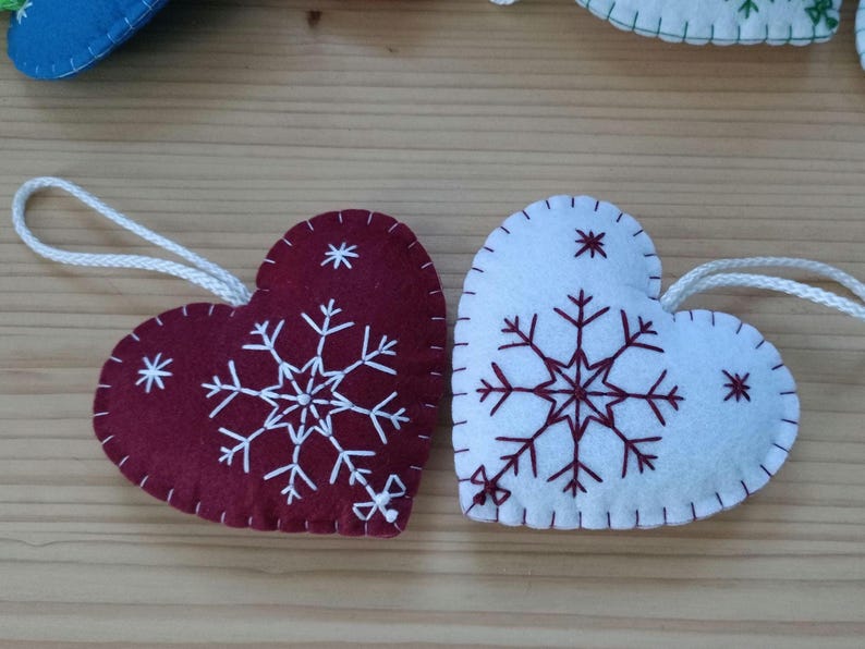 Two heart-shaped Christmas ornaments. One is burgundy with white snowflake embroidery, the other is white with burgundy snowflake embroidery. Both have white string loops for hanging. The ornaments are made of felt.