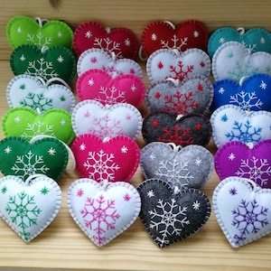 winter and christmas decoration, handmade from felt. twenty four felt hearts with hand embroidered snowflake are set on a display board. these are christmas tree ornaments in wide variety of colors. hearts are plush and with string. lovely.