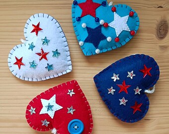 May include: Four felt heart ornaments decorated with red, white, and blue stars and sequins. The hearts are stitched with a white thread. One heart has a blue button in the center.