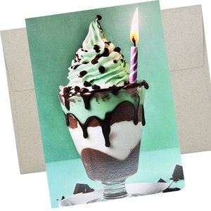 May include: A green mint chocolate chip ice cream sundae with chocolate sauce and a lit candle on top. The sundae is in a clear glass dish with a glass stem and is sitting on a white plate with chocolate shavings.