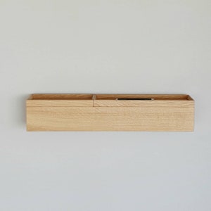 Op de afbeelding: Een licht houten wandorganizer met twee compartimenten. Het linkercompartiment is open, terwijl het rechter een ondiep dienblad heeft met een zwarte pen. De rechthoekige organizer is aan een witte muur bevestigd. Ongeveer 60 cm lang.