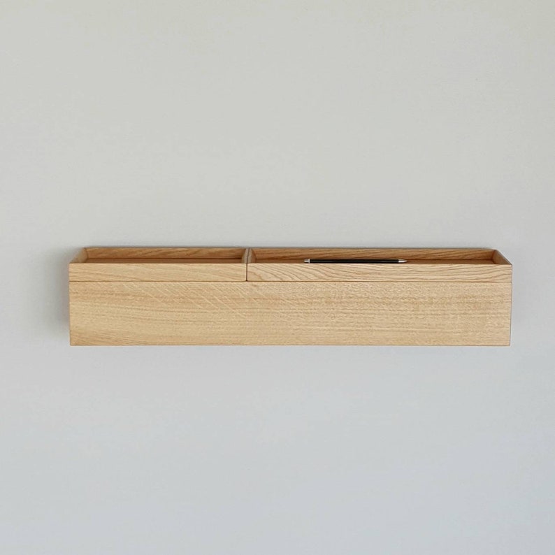 Op de afbeelding: Een licht houten wandorganizer met twee compartimenten. Het linkercompartiment is open, terwijl het rechter een ondiep dienblad heeft met een zwarte pen. De rechthoekige organizer is aan een witte muur bevestigd. Ongeveer 60 cm lang.