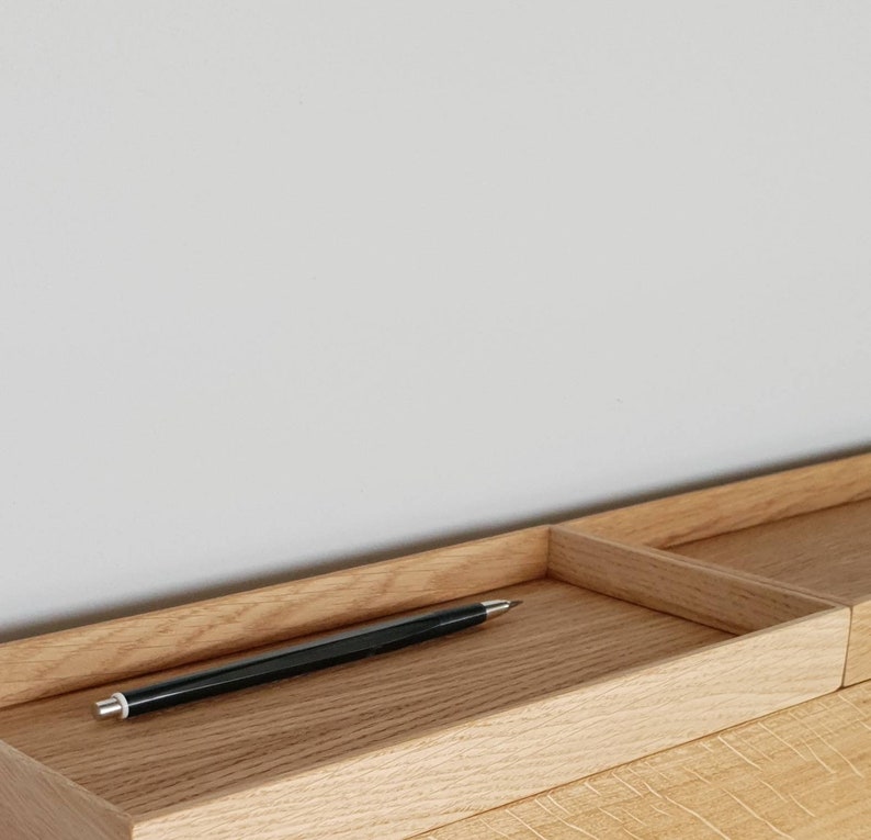 Op de afbeelding: Een lichtgekleurde houten bureau-organizer met een rechthoekige pennenbak en een apart compartiment. Een zwarte pen met een zilveren punt ligt in de bak. De houtnerf is zichtbaar.