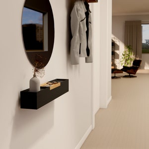 Può includere: Un moderno allestimento per l'ingresso con una mensola nera a parete con un accento in legno, contenente un vaso, fiori secchi e occhiali. Un grande specchio ovale e una giacca colorata completano il design minimalista.