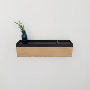 Peut inclure: Étagère murale en bois clair avec un dessus noir. Un petit vase bleu contenant des brins verts est posé sur l'étagère. L'étagère est fixée à un mur blanc, offrant un design minimaliste.