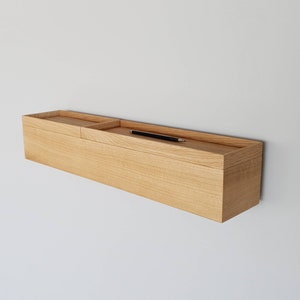 Op de afbeelding: Een licht houten zwevend rek met een verzonken bovendeel en een zwarte pen. Het rek is aan een witte muur bevestigd. Het rek is rechthoekig en lijkt van massief hout te zijn.