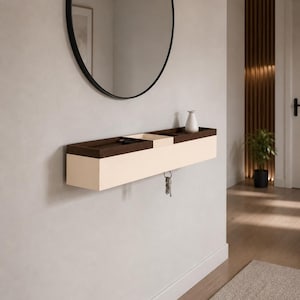 Peut inclure: Une étagère d'entrée murale moderne en crème et marron foncé. Un petit vase blanc est posé sur l'étagère, et des clés sont suspendues en dessous. Un miroir rond noir est accroché au-dessus de l'étagère. L'étagère est fixée à un mur gris clair.