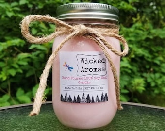 May include: A pink soy wax candle in a clear glass jar with a white label that reads "Wicked Aromas" and "Hand Poured 100% Soy Wax Candle". The label also includes "Made in U.S.A NET WT. 16 oz". The jar is tied with a brown twine bow.