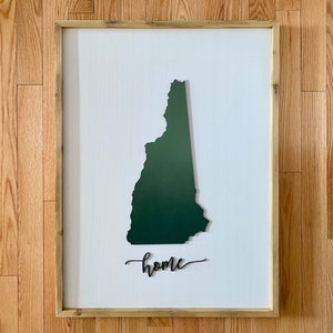 Puede incluir: Una pieza de arte de pared enmarcada que presenta una silueta verde oscura del estado de New Hampshire con la palabra "home" escrita en cursiva debajo.