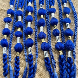 Könnte beinhalten: Geflochtene blaue Haarverlängerungen mit dekorativen blauen Pompons, silbernen und goldenen Akzenten. Die Verlängerungen sind vertikal vor einem neutralen Hintergrund angeordnet. Zöpfe unterschiedlicher Dicke.
