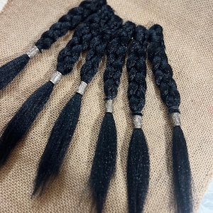 May include: Six black braided hair extensions with silver-colored decorative bands at the base of each braid. The braids are arranged on a tan burlap surface, showcasing the texture and detail of the hair extensions.