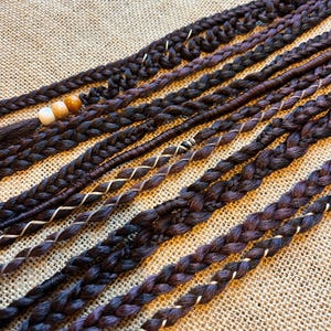May include: Close-up of several dark brown and purple braided hair extensions. Some braids have decorative beads and white or gold accents. The extensions are arranged on a textured, tan surface.