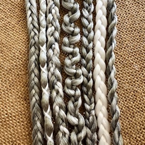 Può includere: Una collezione di otto cordoni intrecciati in varie tonalità di grigio e bianco. I cordoni sono disposti verticalmente e hanno tutti approssimativamente la stessa lunghezza.